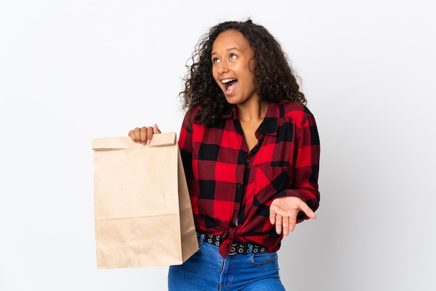 Nastolatek dziewczyna trzyma torbę na zakupy spożywcze na wynos na białym tle na białej ścianie z wyrazem zaskoczenia, patrząc z boku