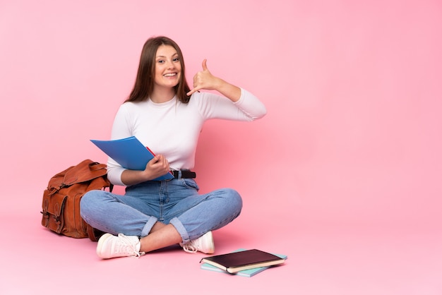 Nastolatek dziewczyny caucasian studencki obsiadanie na podłoga odizolowywającej na menchii ścianie robi telefonu gestowi. Oddzwoń do mnie znak