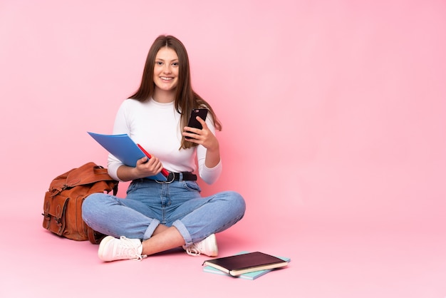 Nastolatek Dziewczyny Caucasian Studencki Obsiadanie Na Podłoga Odizolowywającej Na Menchii ścianie Wysyła Wiadomość Z Wiszącą Ozdobą