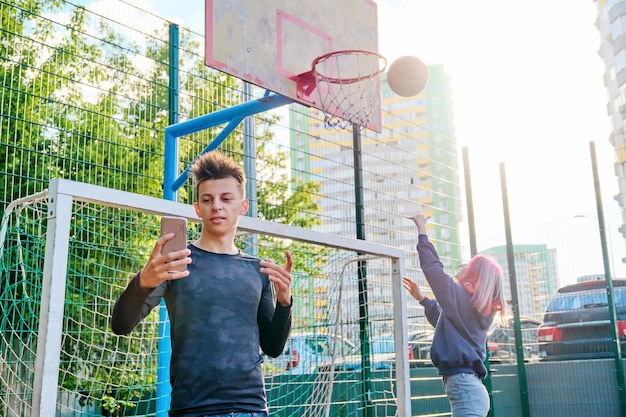 Nastolatek facet nagrywa strumień wideo za pomocą smartfona, na boisku sportowym ulicy, dziewczyna gra w koszykówkę, rzuca piłkę w koszu. Nowoczesna aktywna młodzież, technologia, hobby i wypoczynek