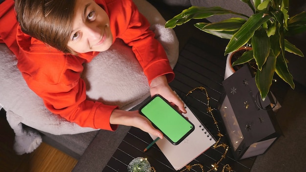 Nastolatek ładny chłopiec w czerwonym swetrze trzymając telefon z zielonym ekranem i patrząc na kamery Wysoki kąt widzenia