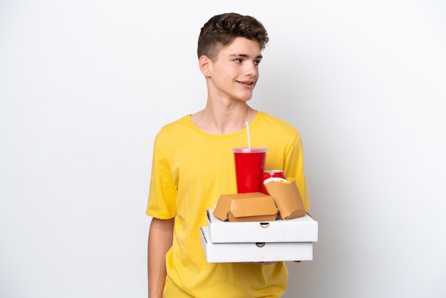 Nastolatek Rosjanin trzymający fast food na białym tle patrząc w bok i uśmiechnięty
