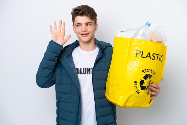 Nastolatek Rosjanin trzymający torbę pełną plastikowych butelek do recyklingu na białym tle słuchający czegoś, kładąc rękę na uchu