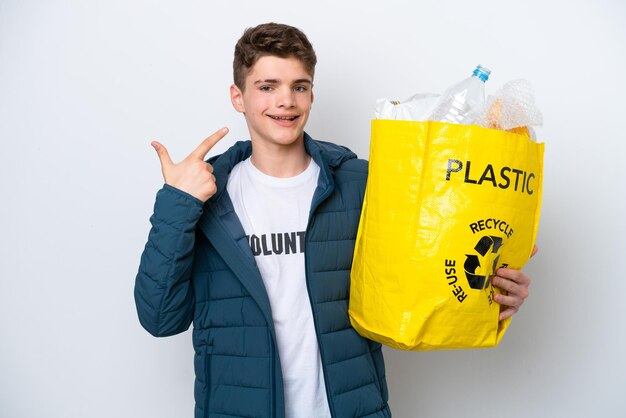 Nastolatek rosyjski trzymający torbę pełną plastikowych butelek do recyklingu na białym tle, pokazując gest kciuka w górę