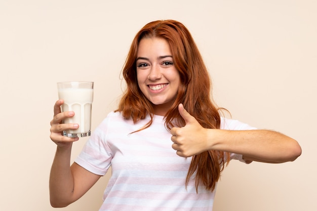 Nastolatek ruda dziewczyna trzyma szklankę mleka na izolowanej ścianie z kciuki do góry, ponieważ stało się coś dobrego