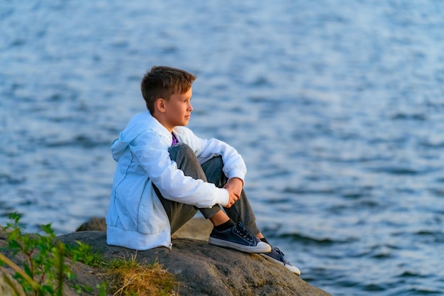 Nastolatek siedzi na dużym kamiennym głazie na brzegu rzeki i patrzy na rzekę poniżej Letnie powołanie Jasnozielona roślinność błękitne niebo