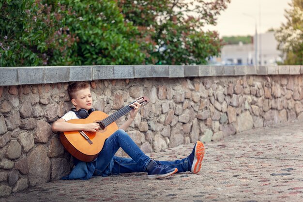 Nastolatek siedzi w parku z gitarą akustyczną i hełmofonami.