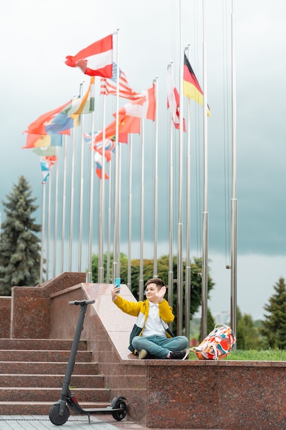 Zdjęcie nastolatek siedzi w pobliżu budynku administracyjnego na tle flag państw i macha do smartfona.