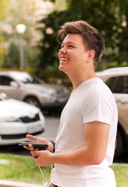 Nastolatek w białej koszulce stoi na ulicy i uśmiecha się, trzymając telefon ze słuchawkami