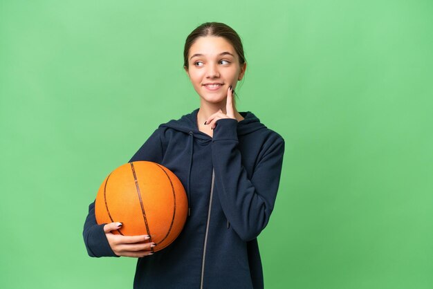 Nastolatka kaukaska dziewczyna gra w koszykówkę na odosobnionym tle, myśląc o pomyśle, patrząc w górę