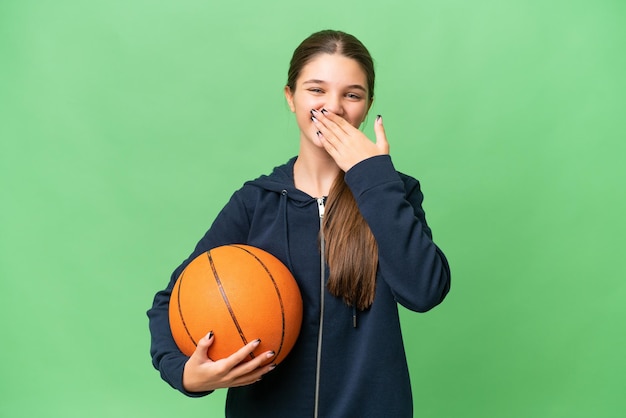 Nastolatka kaukaska dziewczyna gra w koszykówkę na odosobnionym tle szczęśliwa i uśmiechnięta zakrywająca usta ręką