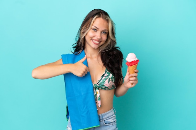 Nastolatka kaukaska dziewczyna trzyma lody i ręcznik na białym tle na niebieskim tle, pokazując gest kciuka w górę