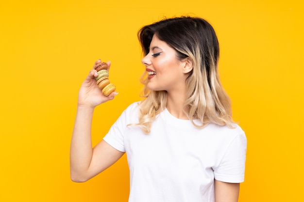 Nastolatka na żółtej ścianie trzymając kolorowe francuskie macarons i szczęśliwy wyrazem twarzy