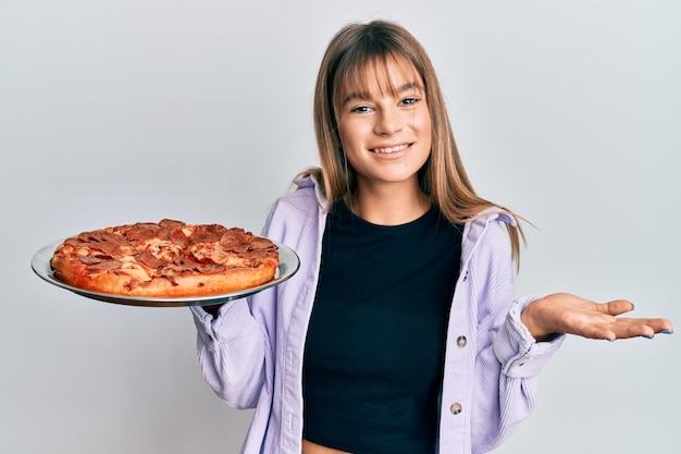 Nastolatka rasy kaukaskiej trzymająca włoską pizzę świętująca osiągnięcie z radosnym uśmiechem i wyrazem twarzy zwycięzcy z podniesioną ręką