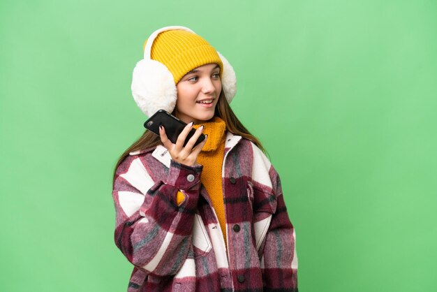 Nastolatka rasy kaukaskiej w zimowych mufkach na odosobnionym tle, prowadząca z kimś rozmowę przez telefon komórkowy