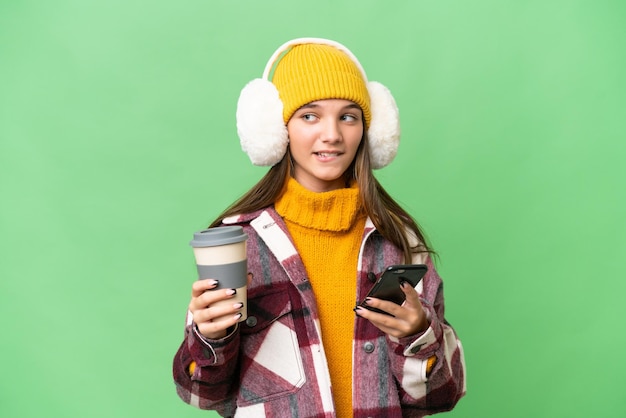 Nastolatka rasy kaukaskiej w zimowych mufkach na odosobnionym tle trzymająca kawę na wynos i telefon komórkowy, myśląc o czymś