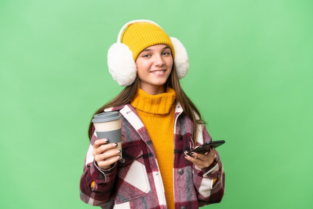 Nastolatka rasy kaukaskiej w zimowych mufkach na odosobnionym tle trzymająca kawę na wynos i telefon komórkowy