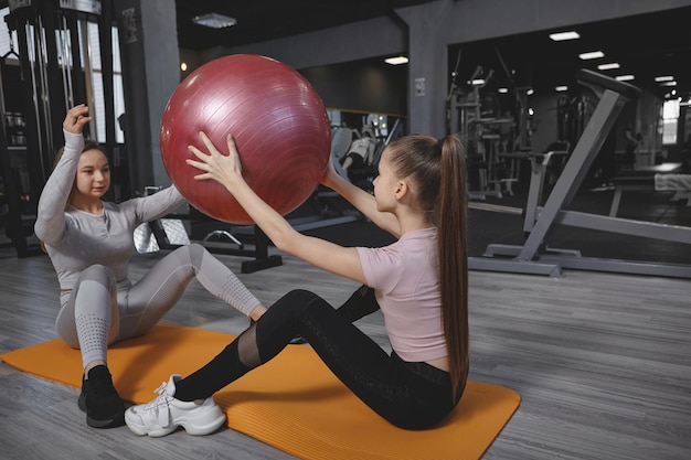 Nastolatka robi przysiady z piłką fitness, ćwicząc z osobistym trenerem na siłowni