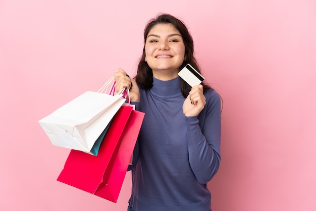 Nastolatka Rosjanka na białym tle na różowej ścianie, trzymając torby na zakupy i kartę kredytową