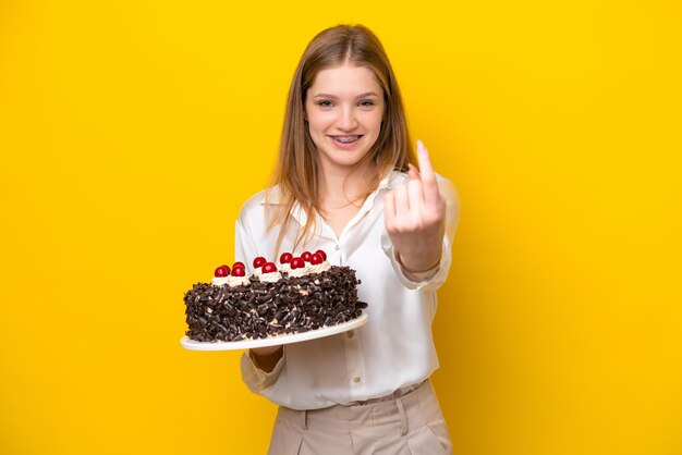 Nastolatka Rosjanka trzyma tort urodzinowy na białym tle na żółtym tle robi nadchodzący gest