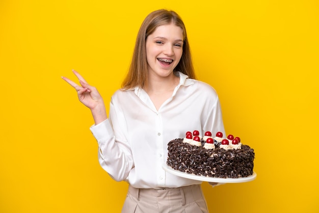 Nastolatka Rosjanka trzyma tort urodzinowy na białym tle na żółtym tle, uśmiechając się i pokazując znak zwycięstwa