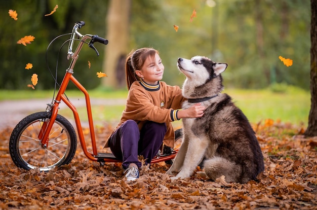 Nastolatka siedzi na skuterze wśród jesiennych liści w parku i przytula swojego psa rasy Siberian Husky