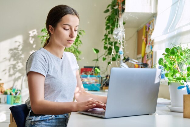 Nastolatka studiująca w domu przy stole za pomocą laptopa Nastoletnia studentka studiująca online odrabiająca pracę domową pisząca w zeszycie oglądająca lekcję wideo Elearning adolescencja technologia w szkole średniej