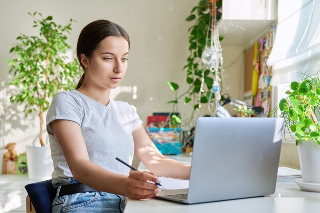 Nastolatka ucząca się w domu przy stole za pomocą laptopa Uczeń nastolatek uczący się online robiąc zadania domowe pisanie w notatniku oglądanie słuchania lekcji wideo Elearning młodzież technologia licealna