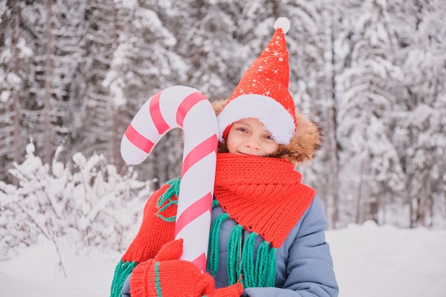 Nastolatka w ciepłym wężowym ubraniu w bożonarodzeniowych kwiatach dziecko na zewnątrz w kapeluszu santa bawi się dużą nadmuchiwaną laską cukrową