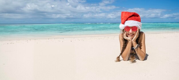 Nastolatka w noworocznym czerwonym kapeluszu i czerwonych okularach przeciwsłonecznych na słonecznej tropikalnej plaży