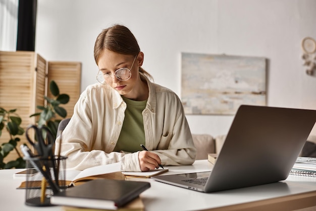 nastolatka w okularach trzymająca długopis i pisząca podczas zajęcia online na laptopie robiąca notatki