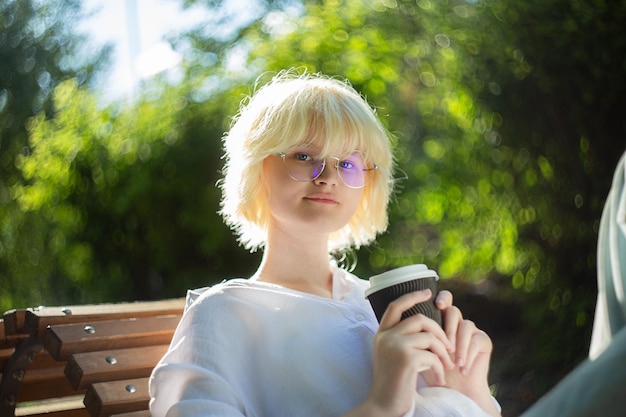 Zdjęcie nastolatka z blond włosami pije kawę z papierowego kubka na zewnątrz.