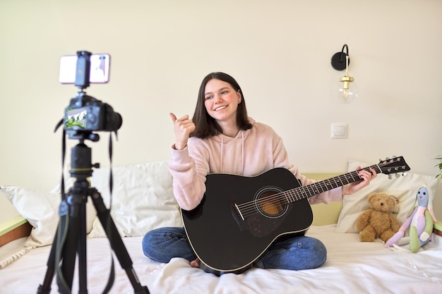 Nastolatka z gitarą akustyczną siedzi w domu na łóżku, nagrywa muzykę na wideo i rozmawia z przyjaciółmi