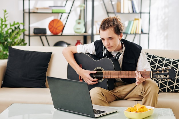 Nastoletni chłopak oglądając seminarium internetowe na komputerze typu tablet i próbując nauczyć się grać na gitarze