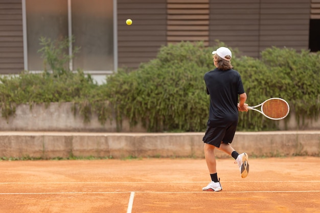 Nastoletni chłopak w czarnej koszulce uderza piłkę tenisową na korcie