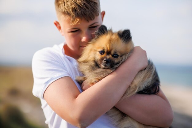 Nastoletni facet w koszulce trzyma w ramionach psa rasy pomorskiej na tle jasnego słonecznego błękitu nieba