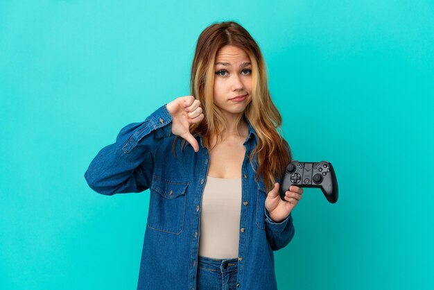 Nastoletnia blondynka bawi się kontrolerem gier wideo nad odosobnioną ścianą pokazując kciuk w dół z negatywną ekspresją