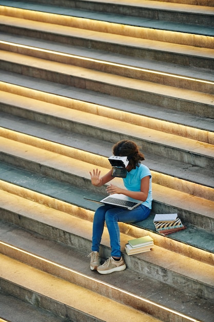 Nastoletnia dziewczyna siedzi na schodach z książkami i laptopem i testuje stworzoną przez siebie aplikację Virtual Geality