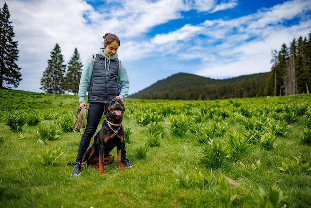 Nastoletnia dziewczyna stoi ze smyczą w rękach obok psa rasy Rottweiler na łące na tle jodły