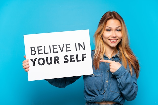Nastoletnia dziewczyna trzyma na białym tle niebieski napis z tekstem „Uwierz w siebie” i wskazując go