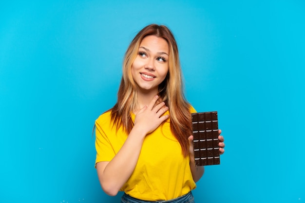 Nastoletnia dziewczyna trzymająca czekoladę na odosobnionym niebieskim tle patrząc w górę podczas uśmiechania się