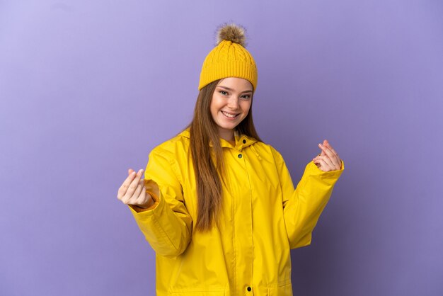 Nastoletnia dziewczyna ubrana w przeciwdeszczowy płaszcz na odosobnionym fioletowym tle robiąca gest pieniędzy