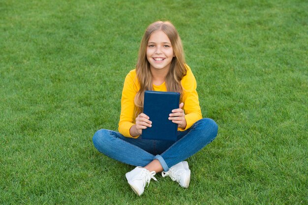 Zdjęcie nastoletnia dziewczyna zainspirowana nową książką. zrelaksować się na zielonej trawie. wiosenny czas wolny. szczęśliwe dzieciństwo. dziecko czytać książki na świeżym powietrzu w parku. szczęśliwa mała dziewczynka studiuje na wakacjach. dziecięca uroda i moda.
