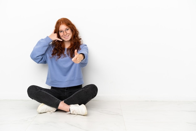 Nastoletnia ruda dziewczyna siedzi na podłodze na białym tle, wykonując gest telefonem i wskazując przód