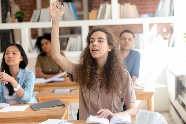 Nastoletnia studentka college'u podnosząca rękę podczas zajęć z wielonarodową grupą kolegów z klasy inteligentna licealistka zadająca pytanie na lekcji siedząca przy biurku koncepcja przywództwa i wiedzy