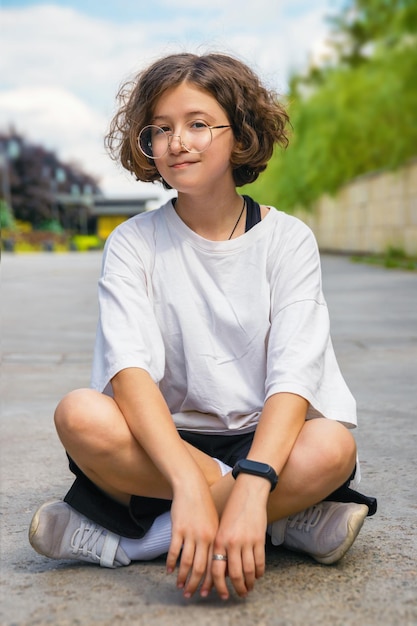Zdjęcie nastoletnie dziewczyny z kręconymi włosami w modnych modnych okularach siedzą na asfalcie w miejskim parku
