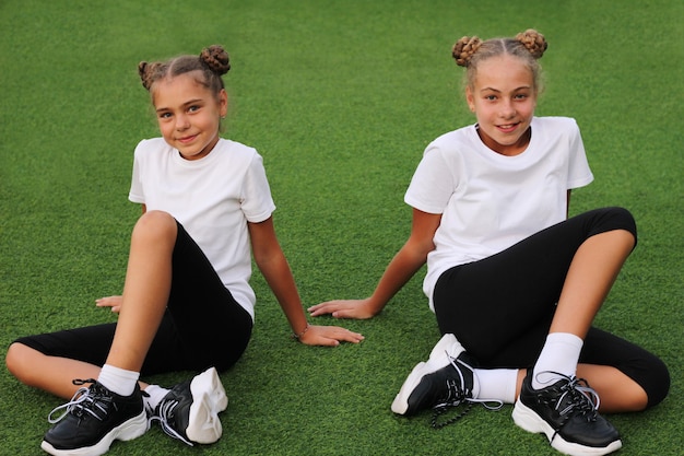 Nastoletnie siostry w strojach sportowych siedzą na zielonej trawie stadionu i patrzą prosto w kamerę. Białe koszulki. Sport w naszym życiu.