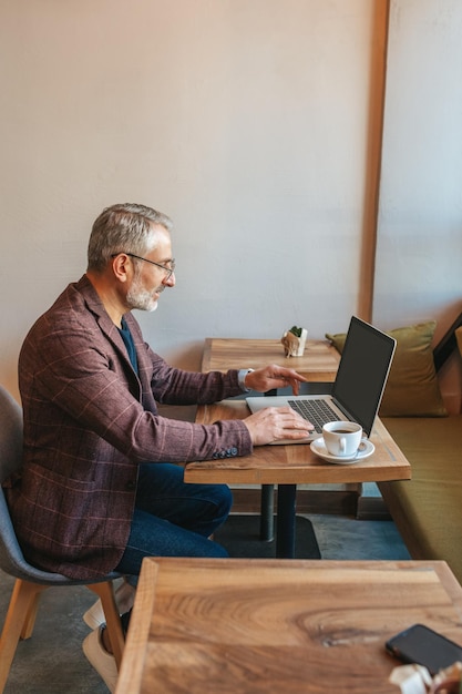 Nastrój pracy. Siwy brodaty elegancki mężczyzna pracujący na laptopie siedzący bokiem do kamery przy stole z poranną kawą w kawiarni