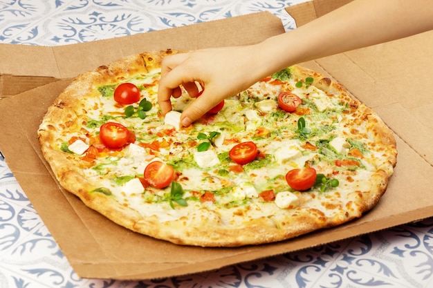Zdjęcie naturalna włoska pizza z dużym pesto z ciastem cienkim kurczak mozzarella pomidory wiśniowe i bazylia w pudełku rzemieślniczym na stole z płytkami ręka dziecka bierze kawałek sera z pizzy kuchni śródziemnomorskiej