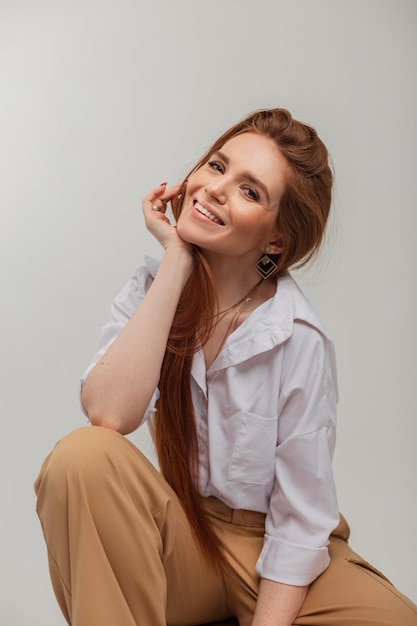 Naturalny kobiecy portret pięknej wesołej, uroczej kobiety z pięknym uśmiechem w modnych ubraniach ze spodniami i koszulą siedzi na białym tle w studio